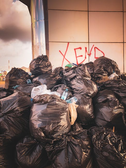 Two large bags of rubbish, made of black plastic and crumpled, are placed on the pavement in front of a black metal fence with vertical bars. The bags appear to contain household waste and are situate